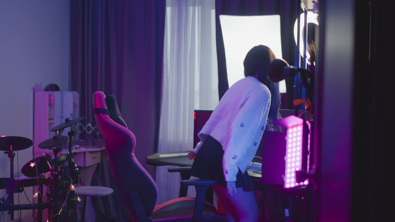 Young girl in fluffy sweater walking into neon lit studio adjusts seat at desk with ring light and white headphone then sits before computer monitors and LED panels under pink purple ambient glow