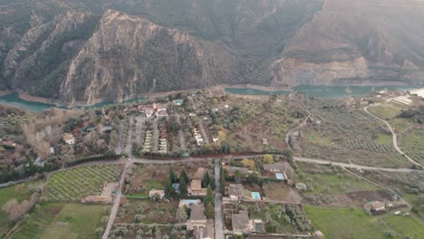 Aerial-view-of-Canales-Reservoir-with-vehicles-moving-on-the-road,-houses,-and-the-big-mountain-behind-,-Spain