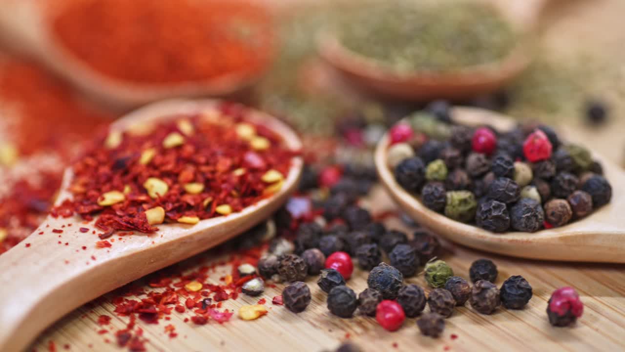 A close-up of various spices on wooden spoons, featuring crushed red pepper flakes and a mix of black, red, and green peppercorns. The spices are scattered on a wooden surface.