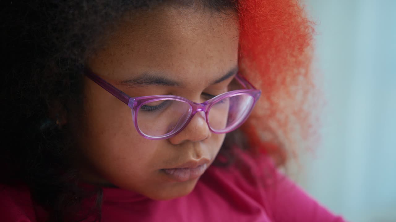 Close up portrait of a young African American girl with purple tinted glasses and partially red hair, engrossed in reading, her facial expressions changing subtly as she processes the information