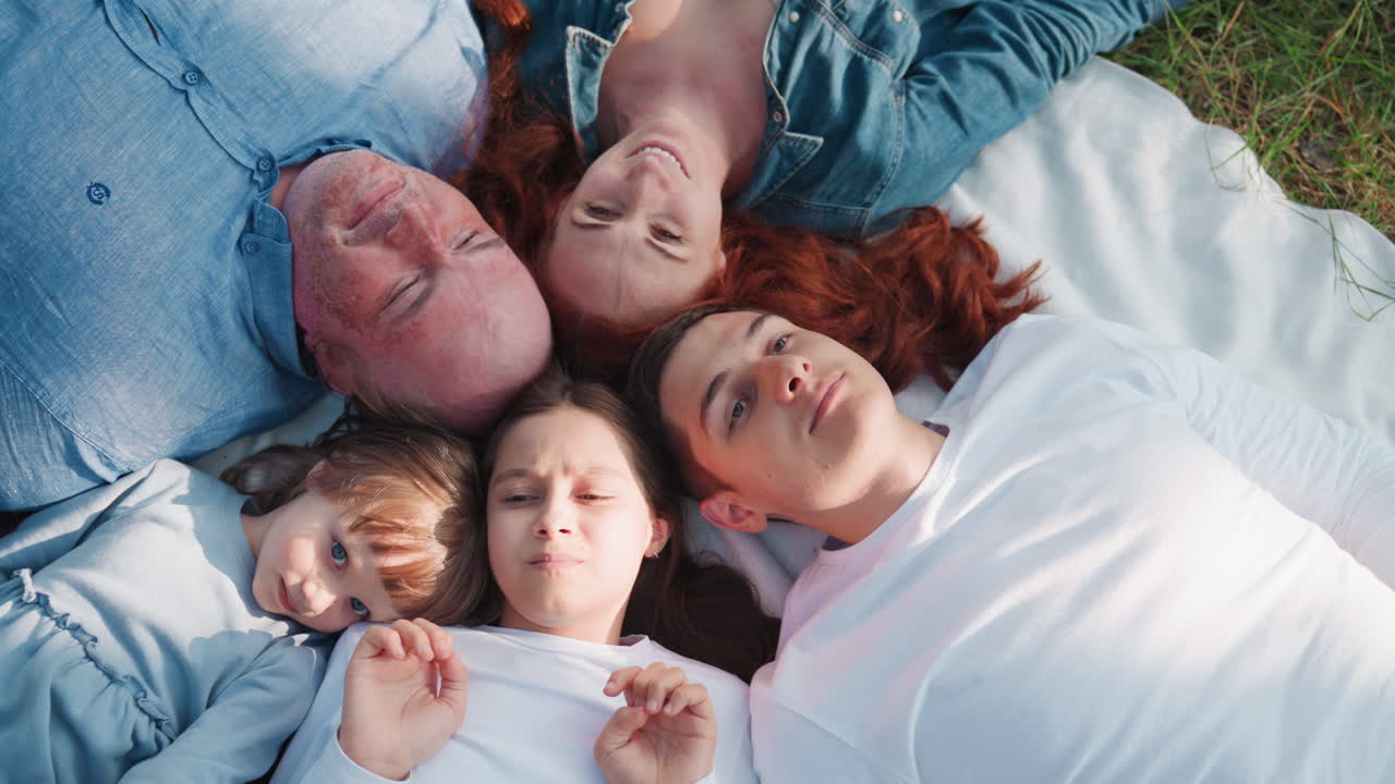 Zoom in on picnic family lying down with heads joined, smiling and talking warmly under sunlight, showing togetherness, love, and harmony in relaxing outdoor atmosphere surrounded by peaceful nature