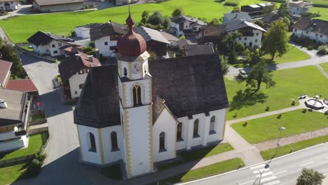 Drone-flying-a-helix-manoever-around-a-scenic-church-in-the-alps-on-a-bright-sunny-day-with-village-and-mountains-in-the-back