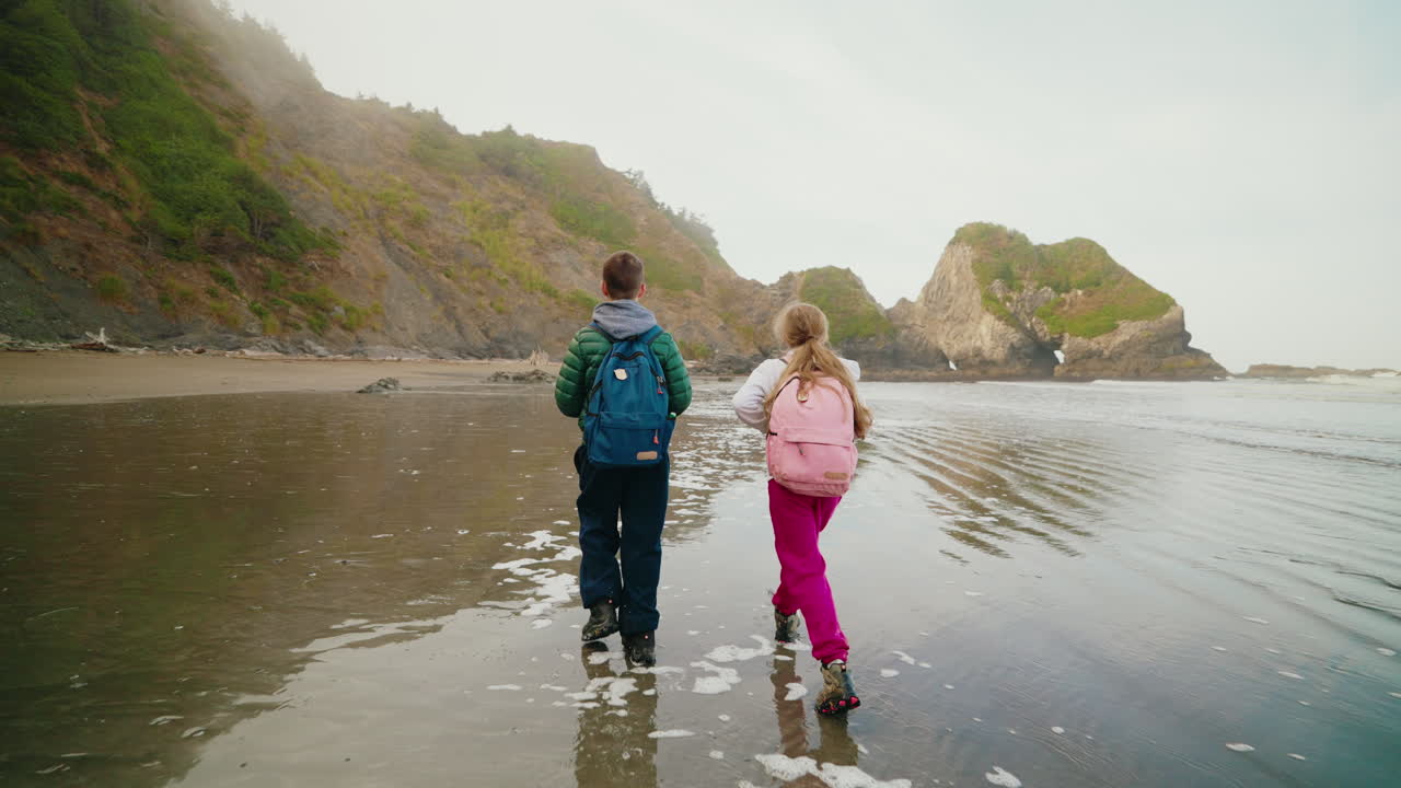 Two Kids with Backpacks Walking on a Scenic Beach Towards Coastal Rock Formations