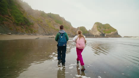 two kids with backpacks walking on a scenic beach towards coastal rock formations