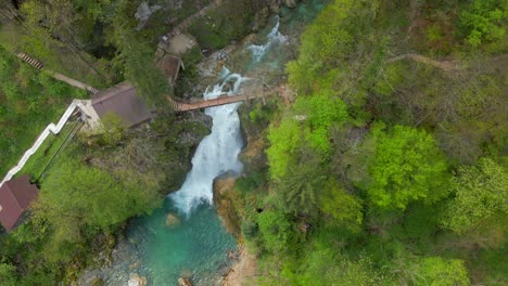 aerial view of a vibrant, turquoise river with a waterfall flowing through lush greenery and a wooden bridge crossing over it