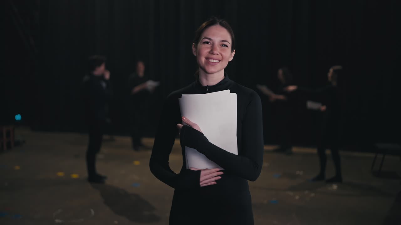 극장에서 무대에서 공연을 준비하는 배우 그룹의 손에 스크립트 종이와 텍스트 한 어리를 가진 검은색 의상을 입은 자신감있는 갈색 배우 소녀의 초상화