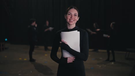 portrait of a confident brunette actor girl in a black suit with a pile of script papers and text in her hands in a group of actors who are preparing to perform on stage in the theater