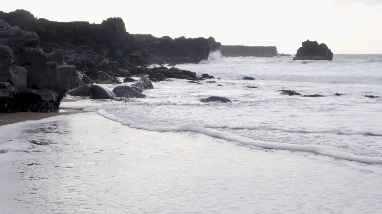 Onde delicate lambiscono una spiaggia di sabbia nera con formazioni di basalto vulcanico al tramonto.