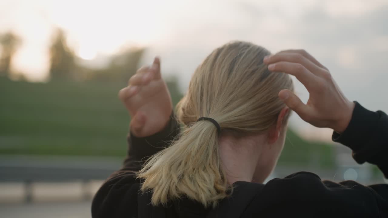 Blond hvit kvinne setter raskt opp hestehale, hender knytter hår og fester strikk, trafikk og bro i bakgrunnen, gyllen time-glød, ryggsekk og hettegenser, rask bevegelse, klar for pendling, cinematisk