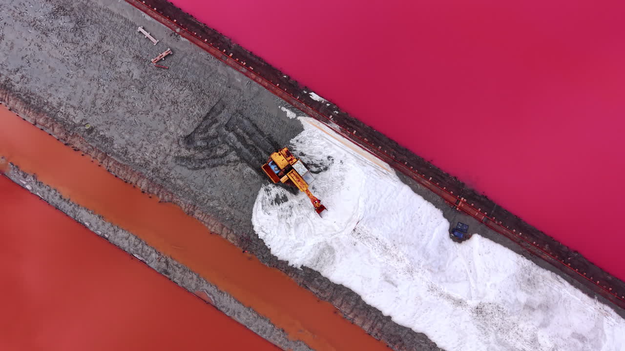Escavatore giallo che sposta sale bianco su una piattaforma delimitata da acqua rosa vibrante. Vista aerea dall'alto di un escavatore che lavora su un cumulo di sale bianco.