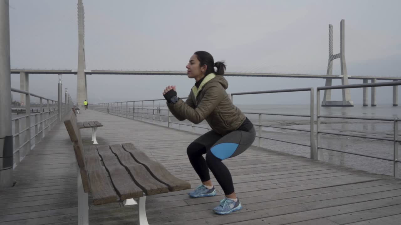 Slow motion shot of concentrated young woman doing squats on wooden pier