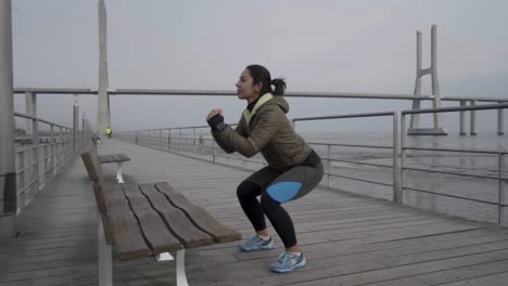 Toma-En-Cámara-Lenta-De-Una-Joven-Concentrada-Haciendo-Sentadillas-En-Un-Muelle-De-Madera