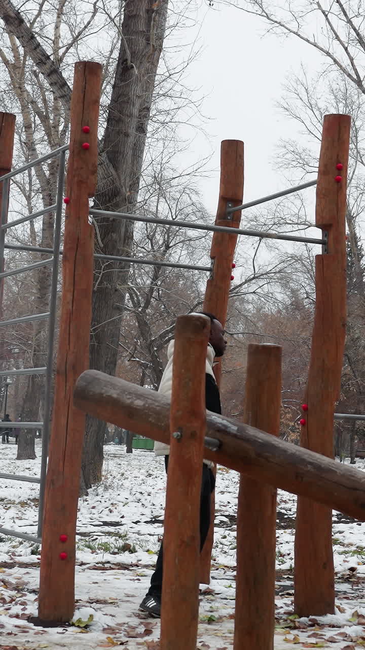 joven adulto participando activamente en flexiones en equipos de entrenamiento al aire libre durante el invierno, rodeado de árboles desnudos en un entorno de parque nevado, ilustrando la dedicación al fitness a pesar del frío