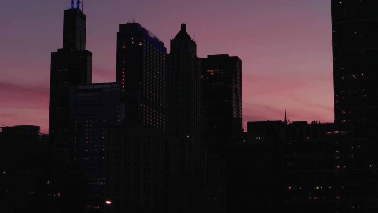 Descenso aéreo del horizonte de Chicago al anochecer, rascacielos silueteados descendiendo hacia el nivel de la calle contra un cielo crepuscular suavemente rosado y morado.