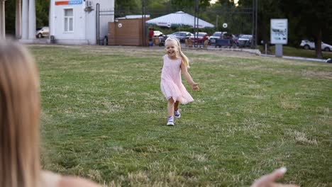 a beautiful blondy girl in a pink dress runs after her mother on the lawn