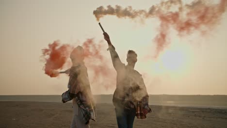 A-happy-brunette-man-with-gray-hair-in-a-checkered-shirt-together-with-his-wife,-a-brunette-girl-in-a-green-checkered-shirt,-holds-fireworks-in-their-hands-that-emit-green-and-red-smoke-during-their-relaxation-and-fun-outside-the-city-in-the-summer-evening