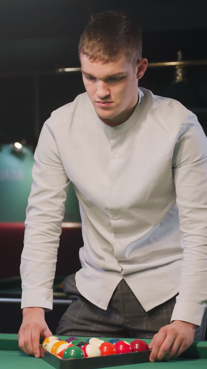 joven con camisa blanca y pantalones grises arregla cuidadosamente bolas de billar de colores en formación de triángulo en una mesa de billar verde vibrante, preparándose para un juego preciso en una sala de billar moderna