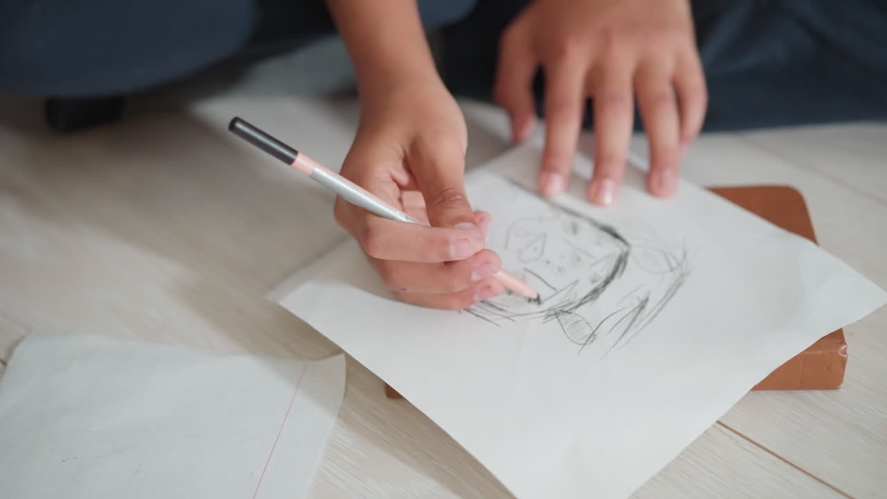 두꺼운 책 표지 위에 놓인 흰 종이에 연필로 스케치하는 아이의 손을 클로즈업한 사진이에요. 단순하고 거친 선과 추상적인 도형으로 창의적인 집중력과 초기 미술 기술을 보여주고 있어요.