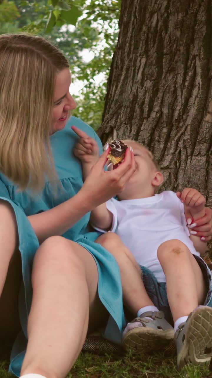 мать с радостью держит своего младшего ребенка, предлагая ему торт, оба улыбаются от радости, старший ребенок смотрит в другую сторону, казалось бы, расстроен