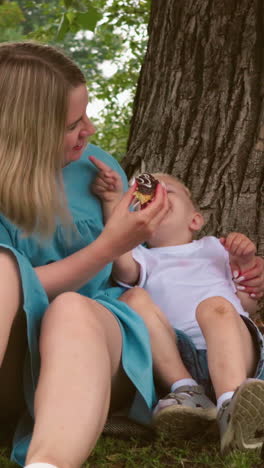 una madre sostiene a su hijo menor felizmente mientras le ofrece un pastel, ambos sonriendo de alegría, el niño mayor mira hacia otro lado, aparentemente molesto