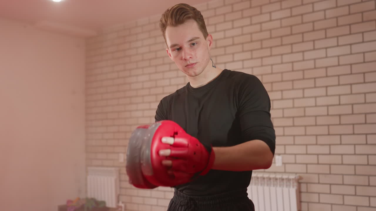 MMA fighter wearing black sportswear prepares for training session holding red boxing pads, focusing on strength, precision, and discipline during practice inside gym with brick wall background and determination