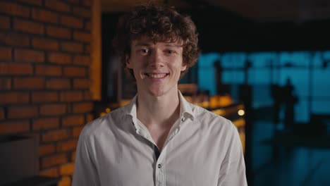 Portrait-of-a-happy-young-guy-in-a-white-shirt-standing-and-posing-in-a-dark-office-with-yellow-lighting