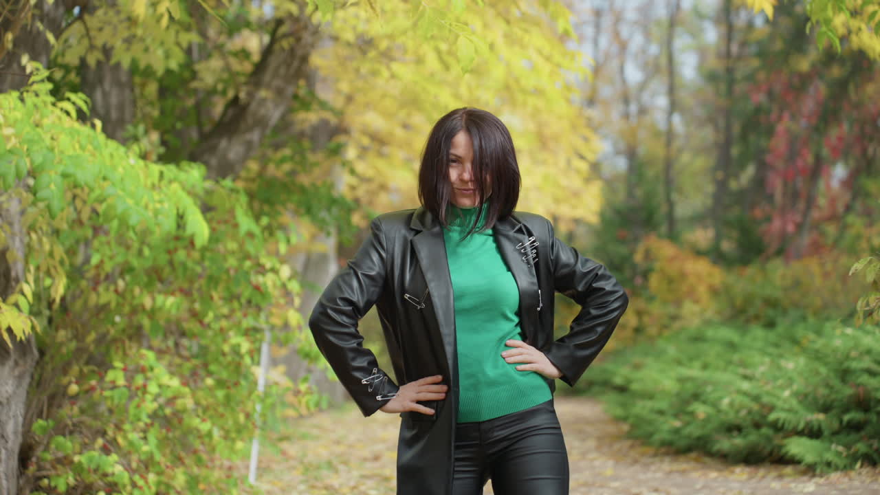Black woman in leather jacket walks along leafy forest path, spins and poses hands on hips, green sweater visible underneath, curly hair framing face, confident rock attitude, boots crunching leaves