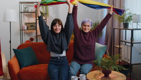 Two-lesbian-women-family-couple-or-girl-friends-holding-LGBT-people-gay-pride-flag-in-hands-at-home