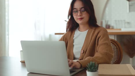 dolly de una mujer asiática segura y alegre abriendo y escribiendo en una computadora portátil. una joven profesional escribiendo un correo electrónico importante desde su casa. sala de estar moderna. trabajo remoto.