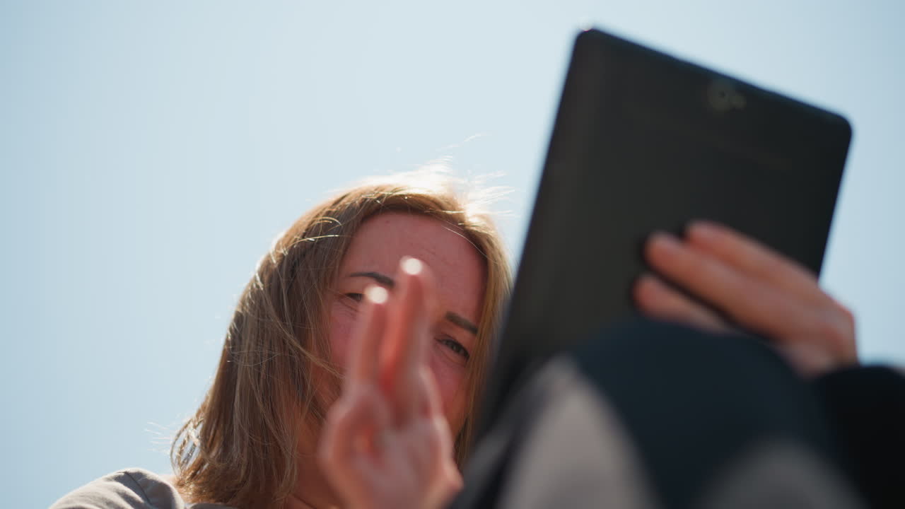 Lady operating phone outdoors looking sad under bright sunlight, wind moving through hair, expression showing emotion and reflection, gentle blur background of trees and sky
