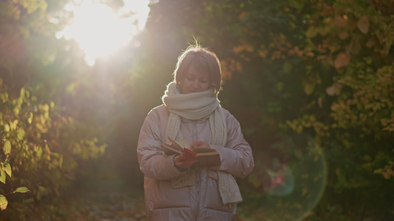 woman walking through autumn garden path flipping book pages in hand under warm sunlight glow around her silhouette, golden foliage backdrop, cozy scarf and jacket