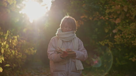 woman walking through autumn garden path flipping book pages in hand under warm sunlight glow around her silhouette, golden foliage backdrop, cozy scarf and jacket