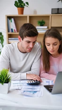 a young couple with a calculator and bills to pay, with a laptop open on the dining table.