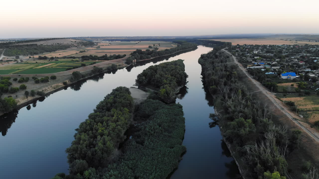 An aerial view showcases a serene river winding through vibrant green fields. The landscape transitions into farmland under a clear twilight sky, highlighting nature's beauty