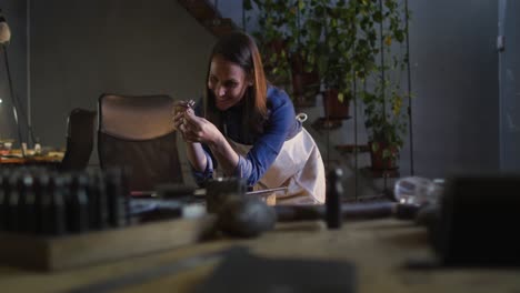 smiling caucasian female jeweller at desk checking quality of jewelry in workshop