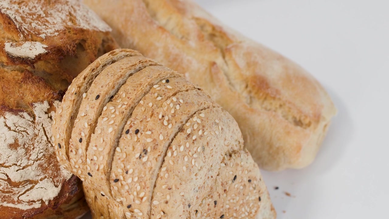 Close-up panning shot of sourdough, sliced loaf, and baguette on a bright white surface.