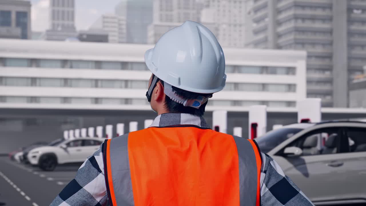 Primo piano posteriore di un ingegnere maschio che indossa un casco di sicurezza, si guarda intorno mentre è in piedi con le mani sui fianchi, veicoli elettrici in carica presso una moderna stazione cittadina