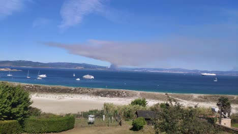 Fuego-En-El-Horizonte-Detrás-De-La-Costa-Con-Huertas-Y-Playa-Con-Yates-De-Lujo-Y-Barcos-Anclados-En-Un-Día-Soleado-De-Verano,-Tiro-Panorámico-Bloqueado,-Islas-Cíes,-Pontevedra,-Galicia,-España