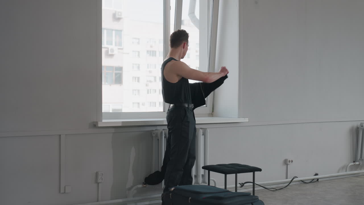Young man standing near window pulls off black jacket, bag rests on floor beside modern stool, high rise office building visible through window, bright minimalist room with neutral walls