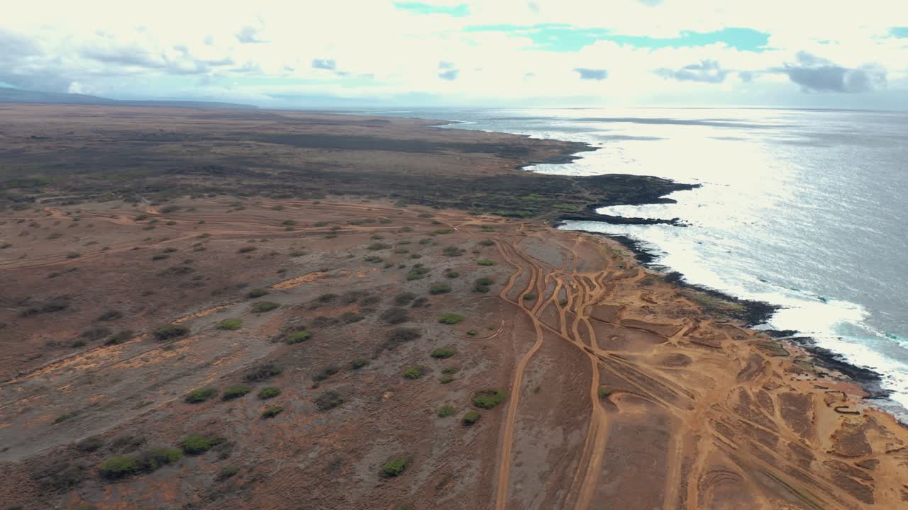A drone glides along an arid coastline where branching trackways descend through scrub to dark rocky shelves, while bright surf flashes under layered clouds, evoking wind, distance, and grit