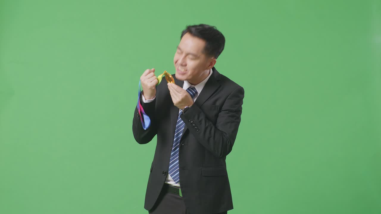 슈트와 타이를 입은 아시아 사업가, 손에 금메달을  자랑스럽게, 스튜디오의 녹색 스크린 배경에서 첫 번째 우승자로 승리합니다.