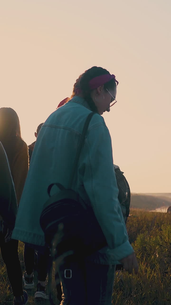 happy young people walk along grass field to cars after day hike against sunset in autumn evening backside view