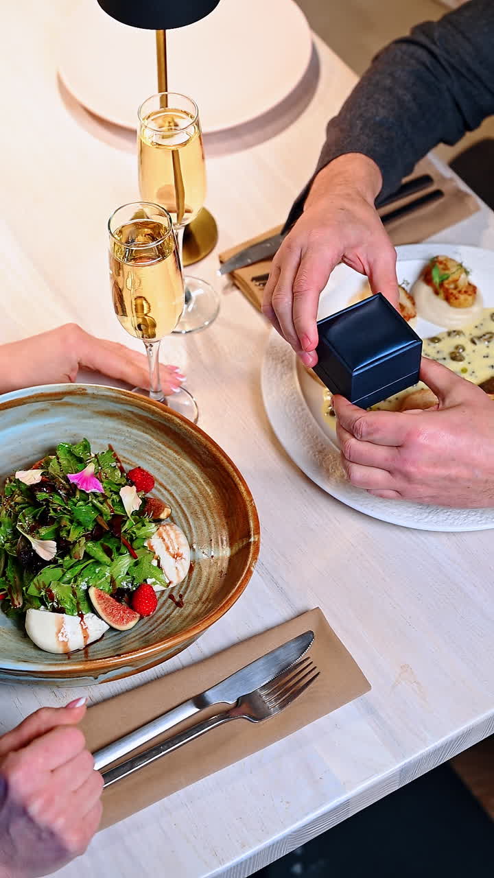 Frieri under middag på en restaurant med elegant mad og funklende drikkevarer. En person overrækker en ring i en æske til en anden ved et spisebord med mad og drikkevarer i baggrunden.