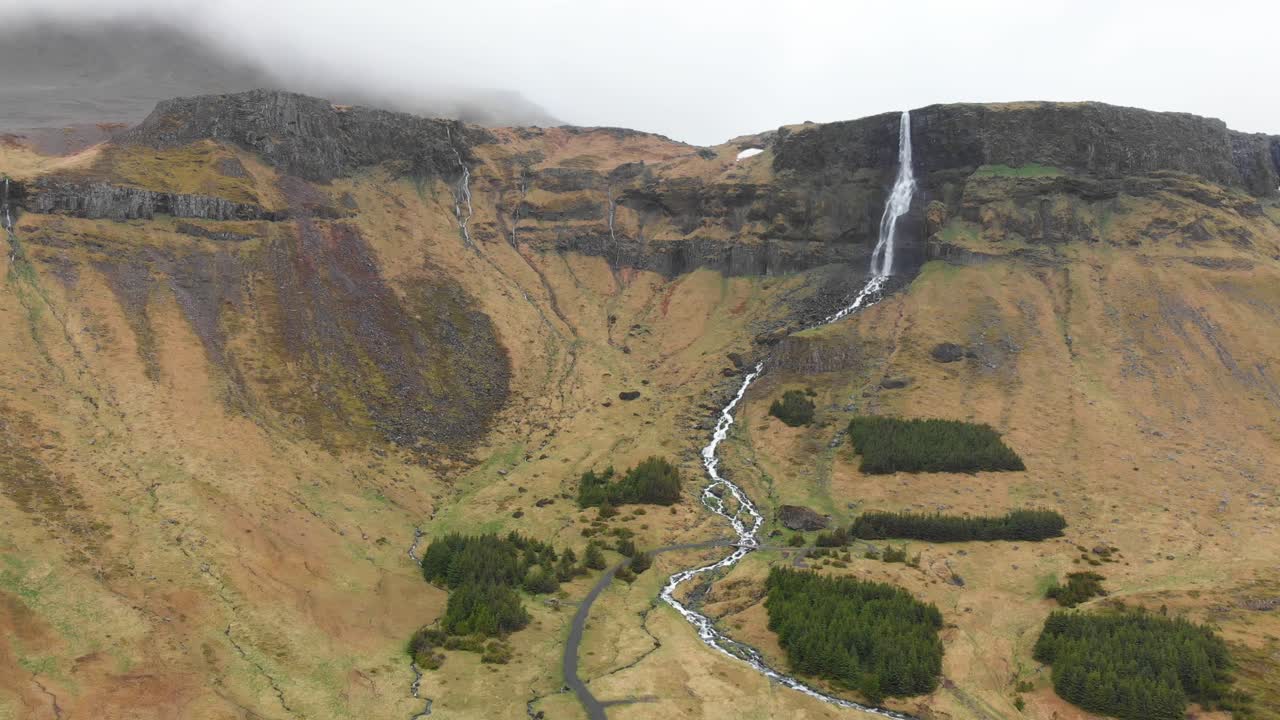 Video aereo delle magnifiche montagne che circondano l'alta e sottile cascata Bjarnarfoss, che sfocia in un fiume nello splendido paesaggio islandese in una giornata nebbiosa e nuvolosa