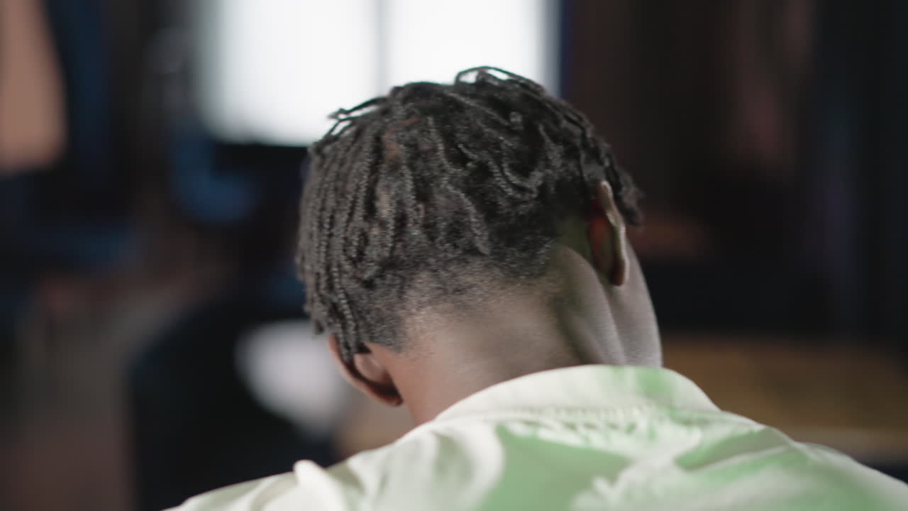 Close-up of a person from behind, showing dreadlocks and white shirt. Background is blurred, with soft light and warm tones. Focus on the back of the head and shoulders in a calm setting