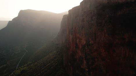 Aerial-push-in-as-light-streams-above-mountain-casting-glowing-rays-to-leonidio-greece-rocky-cliffs