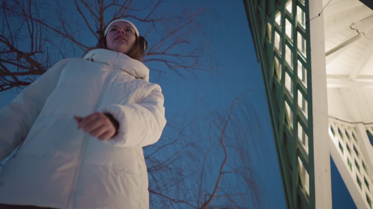 Young lady wearing white puffer coat and headphones spins joyfully under bare winter tree beside illuminated pavilion with snow covered ground and soft evening light highlighting graceful movement