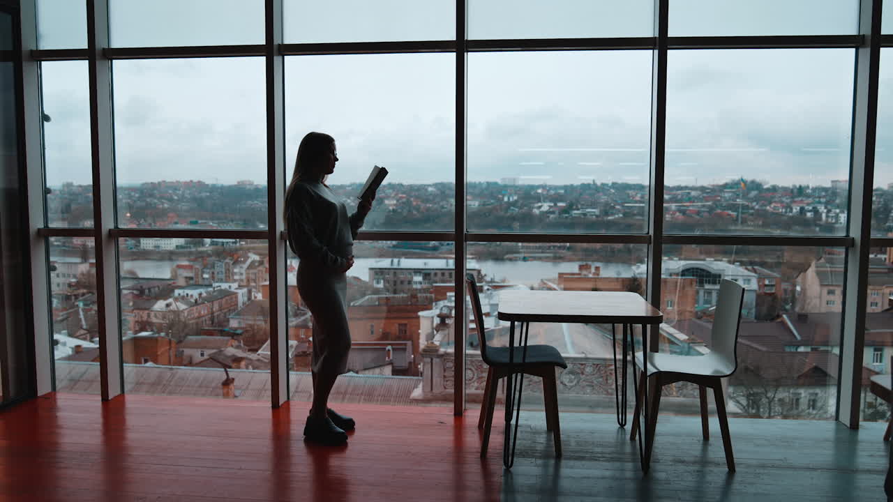 Pregnant woman stands near big window focused on book in her hand. Long-haired lady reads caressing her belly. Cityscape at backdrop.