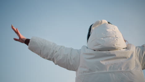 back view of elated young woman twirling joyfully in white winter jacket and beanie, arms raised above head, enjoying calm sunny day with headphones on, under bright blue sky in peaceful open space