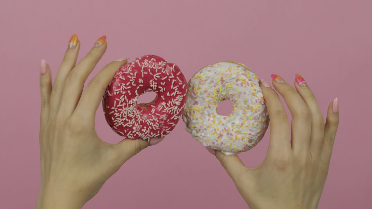 las manos de las mujeres sosteniendo dos rosquillas blancas y rojas, salpicadas sobre un fondo rosa.
